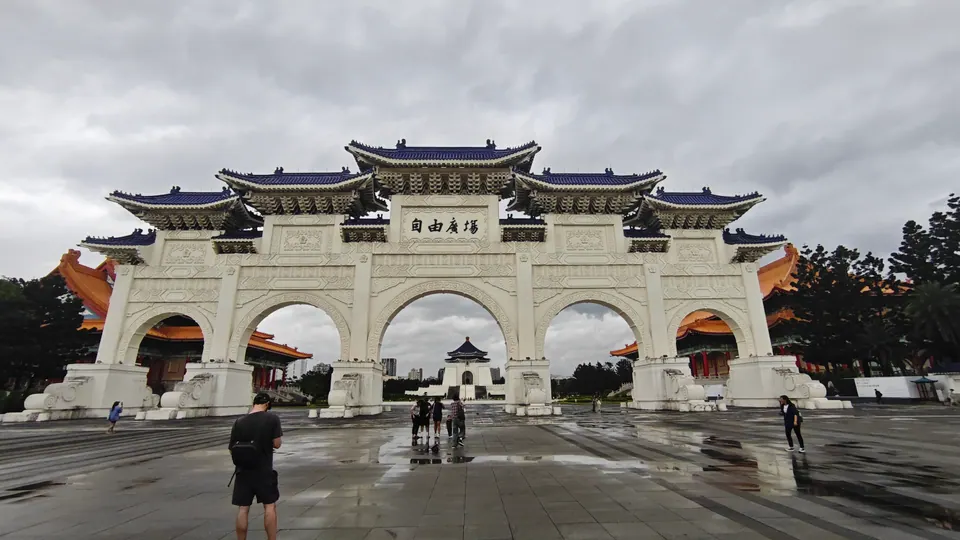 A white gate with blue roofs at Chiang Kai-shek Memorial, overlooking a wet plaza under overcast skies.