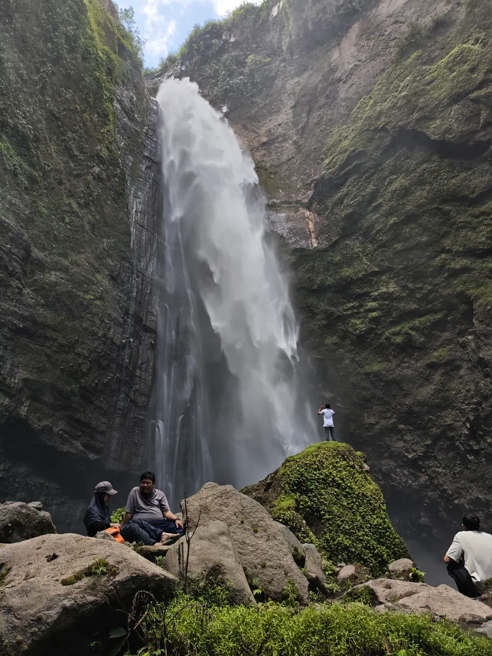 Kabut Pelangi Waterfall flowing between green cliffs, with visitors on the rocks below.